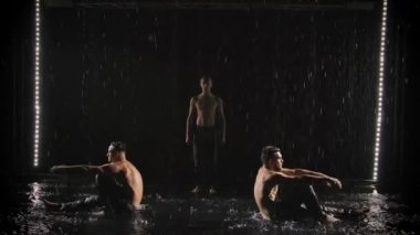 Üç genç sporcu jimnastik dengesi gösteriyor. Erkek akrobatlar. Siyah arka planda, su akıntılarının altında çıplak bir gövde. Yavaş çekim.
