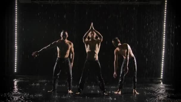 Emocionante espectáculo acrobático bajo la lluvia. Tres hombres posando sobre un fondo de estudio negro retroiluminado en cámara lenta. — Vídeos de Stock
