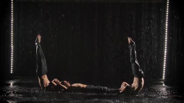 Dos jóvenes atletas demuestran equilibrio gimnástico. Acróbatas macho con un torso desnudo bajo corrientes de agua sobre un fondo negro. Movimiento lento. — Vídeos de Stock