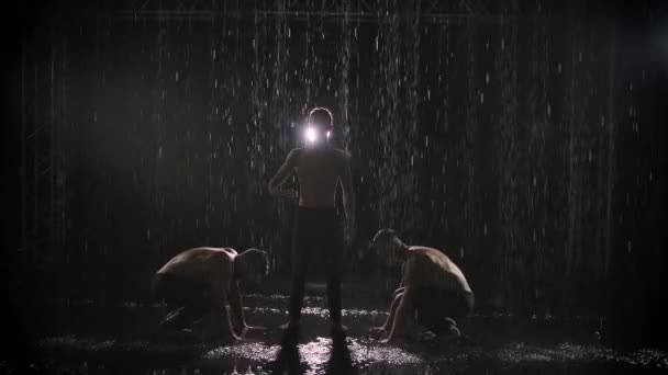 Silhuetas de três homens atléticos realizando acrobacias na chuva. Artistas em destaque em um fundo preto. Movimento lento. — Vídeo de Stock