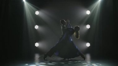 Zarif kıyafetler içinde romantik bir çift vals yapıyor. Sahne ışıkları altında birkaç dansçının siluetleri. Yavaş çekim.