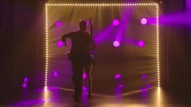 İnce, uzun saçlı bir esmer, partneriyle ça ça dansı yapıyor. Karanlık arka planda parlak ışıkları olan iki kişinin silueti. Yavaş çekim.