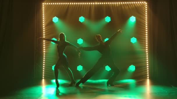 Un par de bailarines profesionales realizan elementos de bailes de salón latinoamericanos rumba, cha cha cha, samba. Siluetas pareja bailando apasionadamente en un estudio oscuro con luces verdes. Movimiento lento. — Vídeo de stock