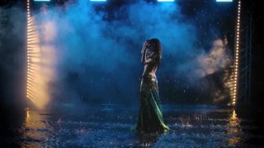 Stüdyoda dumanın arka planına bakan ince saçlı doğulu bir dansçı silueti. Bir kadın suyun yüzeyinde yağmurda dans ediyor. Yavaş çekim.
