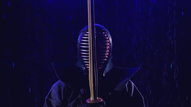 Japon kendosu. Bir kendo savaşçısı zırhlı ve bambu kılıcıyla yağmur damlalarının altında mavi ışıkla aydınlatılmış karanlık bir stüdyonun arka planında duruyor. Kapatın. Yavaş çekim.