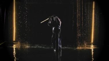 Zırh ve geleneksel kimono giyen bir Japon kendo dövüşçüsü, yağmurda siyah bir zemine karşı bambu bokken ile dövüş sanatı icra ediyor. Yavaş çekim.