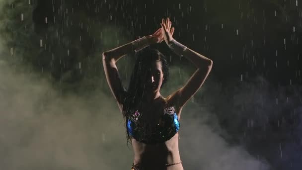 Sylwetka orientalnej urody tańczącej taniec brzucha w deszczu. Kobieta z atrakcyjnym ciałem błyszczącym od kropel deszczu na dymiącym tle studia w zwolnionym tempie. Zamknij się.. — Wideo stockowe