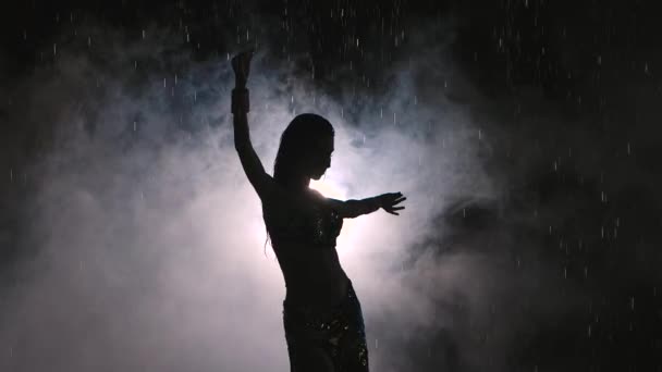 Silhouette scura di ballerino esotico su uno sfondo fumoso in uno studio buio. Una donna balla seducente tra le gocce di pioggia in un abito orientale. Al rallentatore. Da vicino.. — Video Stock