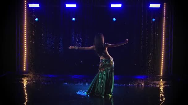 La bruna sta ballando una danza del ventre in un vestito esotico lucido tra le gocce di pioggia. Una donna muove eroticamente i fianchi in uno studio buio e retroilluminato. Rallentatore. — Video Stock