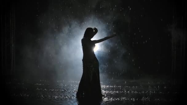 Dark silhouette of a belly dancer on a smoky background among raindrops. Young woman erotically moves her body in the spotlight. Slow motion. — Stock Video