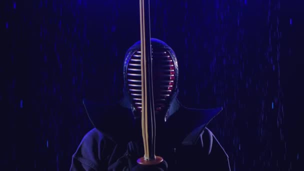 Kendo japonés. Un luchador de kendo está en armadura y con una espada de bambú contra el fondo de un estudio oscuro iluminado por la luz azul bajo las gotas de lluvia. De cerca. Movimiento lento. — Vídeos de Stock