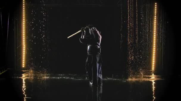 A Japanese kendo fighter wearing armor and traditional kimono practices a martial art with a bamboo bokken against a black background in the rain. Slow motion. — Stock Video