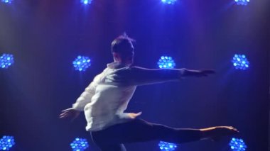 Modern bale yapan yakışıklı genç adam, piruet yapıyor. Koyu dumanlı bir stüdyoda mavi ışıkla dans eden bir adamın silueti. Ağır çekimde kapat..