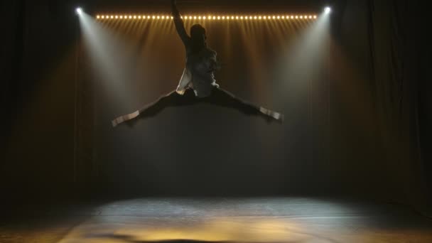 Současný balet, muž provádí prvky moderní choreografie a skoky ve světle reflektorů. Siluety mužské tanečnice v tmavém studiu. Zpomalený pohyb. — Stock video