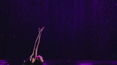Suyun yüzeyinde dans ederek vücudunun esnekliğini gösteren siluet ince jimnastikçi. Sporcu kadın, yağmurda karanlık bir stüdyoda performans sergiliyor. Ağır çekimde kapat..