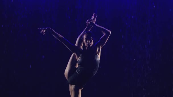 Silhouet slanke turnster die de flexibiliteit van haar lichaam demonstreert door te dansen op het wateroppervlak. De sportvrouw treedt op in de regen op een donkere studioachtergrond. Sluiten in slow motion. — Stockvideo