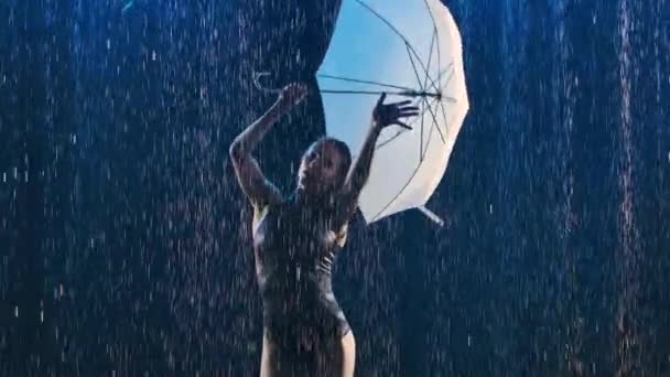 Gimnasta rítmica bailando con un paraguas bajo gotas de lluvia en un estudio oscuro con contraluz azul. De cerca. Movimiento lento. — Vídeos de Stock