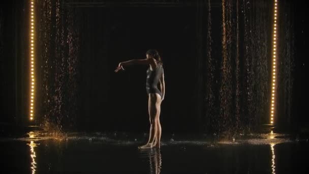 Pesenam rapuh melakukan gerakan akrobatik di permukaan air dalam gerakan lambat. Womans langsing wet body moves in a dark studio with back light. — Stok Video