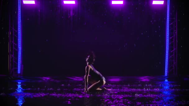 Une jeune athlète féminine fait preuve d'éléments de gymnastique rythmique et d'acrobatie sous les cours d'eau. Silhouette sur fond sombre avec lumière violette au ralenti. — Video