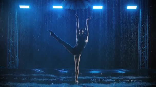 暗いスタジオで雨の流れの下で傘とアクロバティックなパフォーマンス。体操選手はスローモーションで水面で踊っています. — ストック動画