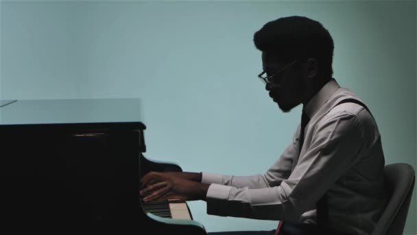 Jonge aantrekkelijke Afro-Amerikaanse pianist in overhemd en stropdas speelt op een prachtige zwarte vleugel. Neergeschoten op een grijze achtergrond met zwak licht, close-up. Langzame beweging. — Stockvideo