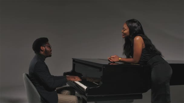 Um homem em um terno listrado elegante canta e toca piano de cauda preto para sua namorada. Feliz casal afro-americano gosta de música. Movimento lento. Fechar. — Vídeo de Stock