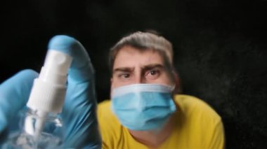 Tıbbi maske takan ve koruyucu eldiven giyen bir adam antibakteriyel spreyi kameraya sıkar ve yüzeyi temizler. Siyah bir arkaplanda çekilmiş, yavaş çekimde kapatılmış..