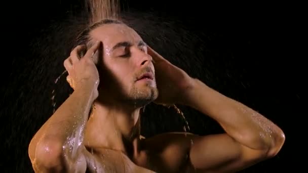 Attraktiver muskulöser junger Mann genießt eine Dusche mit geschlossenen Augen auf schwarzem Hintergrund. Wasserstrahlen fließen in Zeitlupe über nackte Schultern und Arme in Nahaufnahme. — Stockvideo