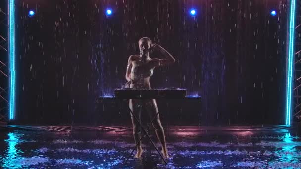 Loira sexy DJ dançando e tocando atrás de baralhos em um estúdio escuro sob chuva e luz azul. Um corpo nu com uma pintura prateada brilha nas gotas de chuva. Movimento lento. — Vídeo de Stock