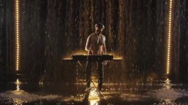 Çıplak göğüslü bir erkek DJ pikaplarda elektronik dans müziği çalar ve karanlık bir stüdyoda yağmurdan zevk alır. Yavaş çekimde karanlık bir arkaplan karşısında video çekmek.