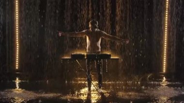 Çıplak göğüslü bir erkek DJ pikaplarda elektronik dans müziği çalar ve karanlık bir stüdyoda yağmurdan zevk alır. Yavaş çekimde karanlık bir arkaplan karşısında video çekmek.