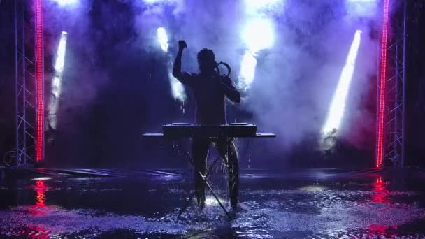 Atractivo dj masculino bailando y tocando música en un estudio oscuro con luces azules y humo. Silueta de un hombre con un torso desnudo, brillante en gotas de lluvia. Movimiento lento. — Vídeos de Stock