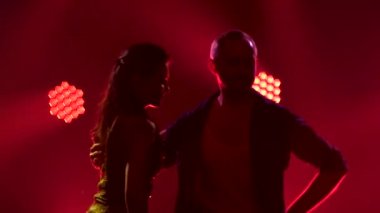 Salsa uzmanlarının siluetleri. Siyahlı genç bir adam parlak kısa elbiseli güzel bir kadınla dans ediyor. Stüdyoda sigara iç. Dinamik çok renkli neon ışıklar. Ağır çekim. Kapat..