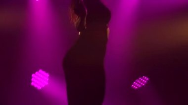 Seksi esmer, erotik olarak dans ediyor ve bükülüyor. İnce bir vücudun silueti stüdyo ışıklarının arka planına yakın. Yavaş çekim.