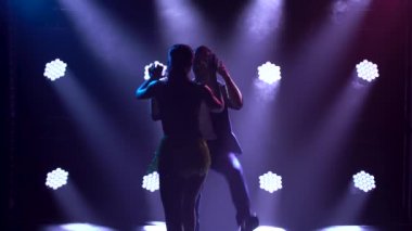 Birkaç profesyonel balo dansçısı Latin müziği eşliğinde dans ediyor ve kalçalarını tutkuyla oynatıyorlar. Bachata, salsa, rumba. Arka planda mor neon ışıkları olan karanlık dumanlı bir stüdyoda yakın çekim..