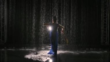 Yağmurda Arjantin flamenkosu dansı yapan ince bir bayan dansçının silueti. Ellerin zarif hareketleri ve ayakların yavaş çekimde sıçraması. Arkaplan ışıklı siyah arkaplan.