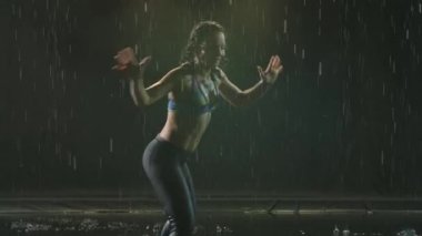Enerjik neşeli kadın dumanlı karanlık stüdyo arka planında capoeira çalışıyor. Sporcu, yağmurda suyun yüzeyinde antrenman yapar ve bir sürü sıçrama yaratır. Siluet. Yavaş çekim. Yakın çekim.