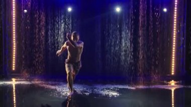 Kaslı bir capoeira akrobat sporcusu yağmurda bir sürü su sıçraması yaratarak numaralar yapar. Koyu arkaplan ve mavi arka ışık. Sağlıklı yaşam tarzı, spor aşkı. Siluet. Yavaş