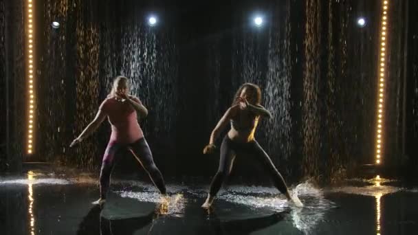 Dos mujeres en forma realizan intrincados movimientos de artes marciales de capoeira al atardecer bajo la lluvia. Parkour acrobático en la superficie del agua con muchas salpicaduras. Silueta. Movimiento lento. — Vídeos de Stock