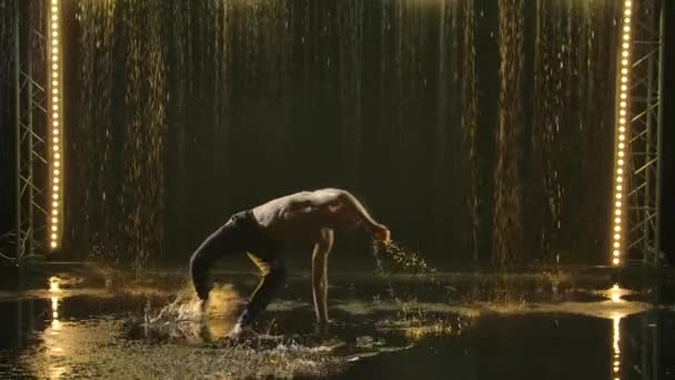 Ein muskulöser Capoeira-Akrobat führt im strömenden Regen Kunststücke vor, die für jede Menge Wasserspritzer sorgen. Dunkler Hintergrund mit Gegenlicht. Gesunder Lebensstil, Liebe zum Sport. Silhouette. Zeitlupe. — Stockvideo