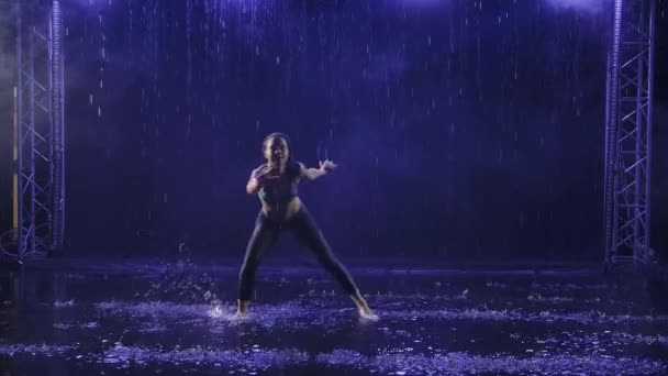 Sylwetka wysportowanej kobiety, pokazująca technikę sztuki walki capoeira w promieniach niebieskiego światła pod strumieniami deszczu. Zwolniony ruch. — Wideo stockowe