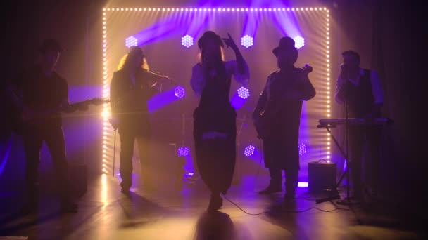 Mladá sólistka zpívá do mikrofonu, když stojí na pódiu s hudební skupinou. Rocková kapela v kostýmech irského stylu proti tmavému studiovému pozadí a modrým světlům. Siluety. Zpomalený pohyb — Stock video
