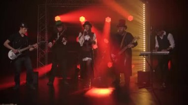 Karanlık stüdyoda müzik performansı sırasında rock grubunda genç bayan şarkıcı ve dinamik renkli ışıklar. Şapkalı grup üyeleri elektro gitar, keman, sentezleyici ve davul çalıyorlar. Yavaş çekim.