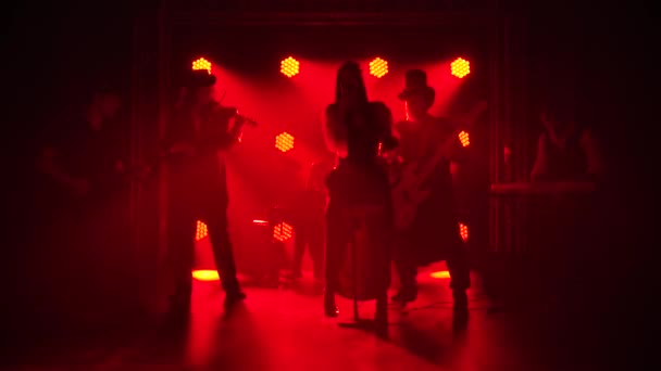 Músicos da banda de rock no estúdio tocam músicas com luzes vermelhas no fundo. Cantora feminina elegante canta com um microfone e os homens tocam instrumentos. Silhuetas. Movimento lento. — Vídeo de Stock