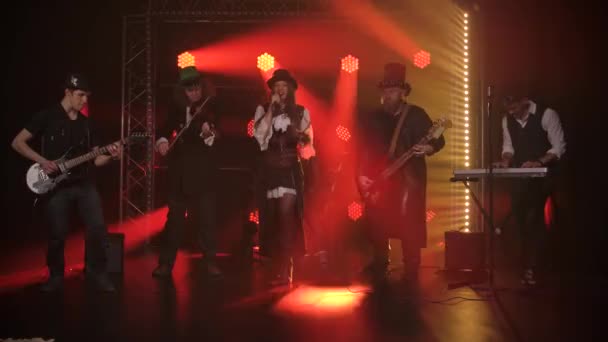 Young woman singer with rock band during music performance in dark studio with dynamic colored lights. Group men in hats are playing the electric guitar, violin and synthesizer and drums. Slow motion. — Stock Video