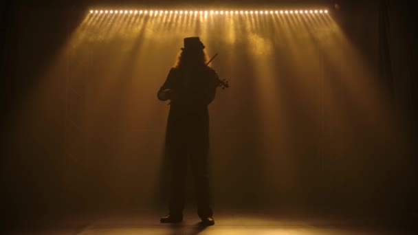 Ein schillernder Geiger mit üppigem Haar und Hut spielt die klassische Holzgeige. Gedreht in einem dunklen, verrauchten Studio mit Lichtern in Zeitlupe. — Stockvideo