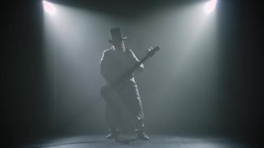 Sakallı rock gitaristi bir stüdyo gösterisinde duman ve neon ışıklarla solo bir rol oynar. Şapkalı ve uzun pelerinli bir adamın silueti yavaş çekimde gitar çalıyor..
