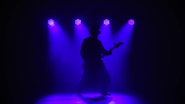 A dark silhouette of a man on a blue background plays the electric guitar. Male musician in a long cloak and hat plays rock music in the studio. Live show.