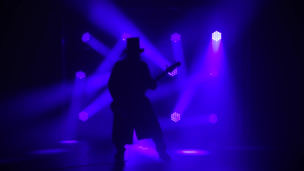 Las siluetas de un hombre sobre un fondo azul toca la guitarra eléctrica. Músico masculino con un impermeable largo y un sombrero al estilo irlandés toca música rock en el estudio. Show en vivo. — Vídeo de stock
