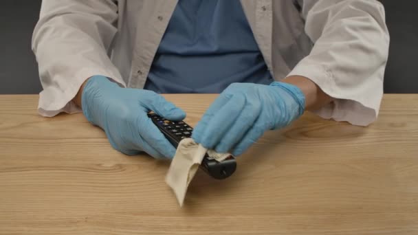 青の保護手袋と白い医療用コートの男は、デバイスをきれいにするために消毒性抗菌ワイプを使用して黒のテレビのリモコンをきれいにします。Covid-19消毒コンセプト。閉めろ。スローモーション. — ストック動画